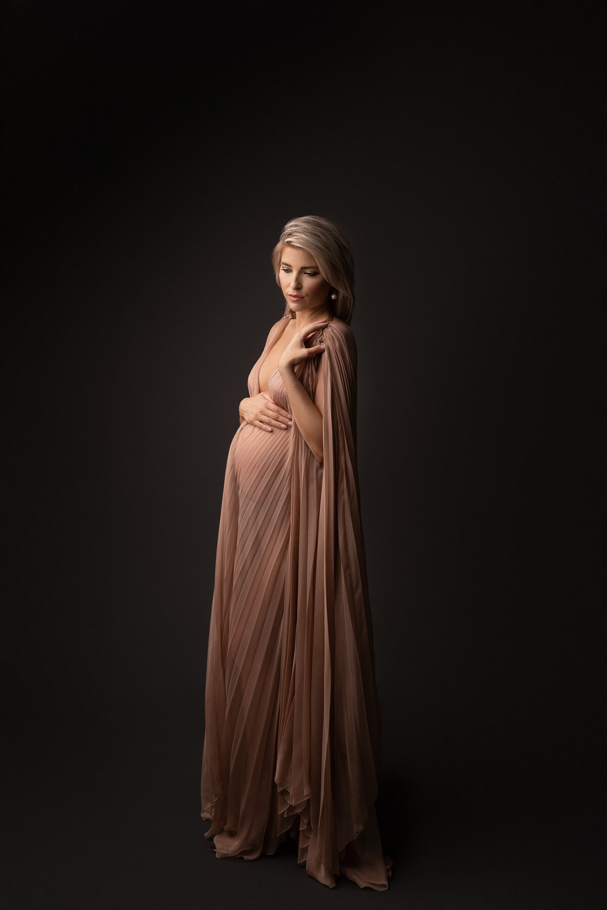 Woman in long dusty rose organza maternity sheath with cape poses for New Jersey maternity photoshoot. She is angled away from the camera with her back arm resting atop of her baby bump and  her forearm is touching her shoulder. She is looking down gently toward her baby bump. Captured by best NJ maternity photographer Katie Marshall.