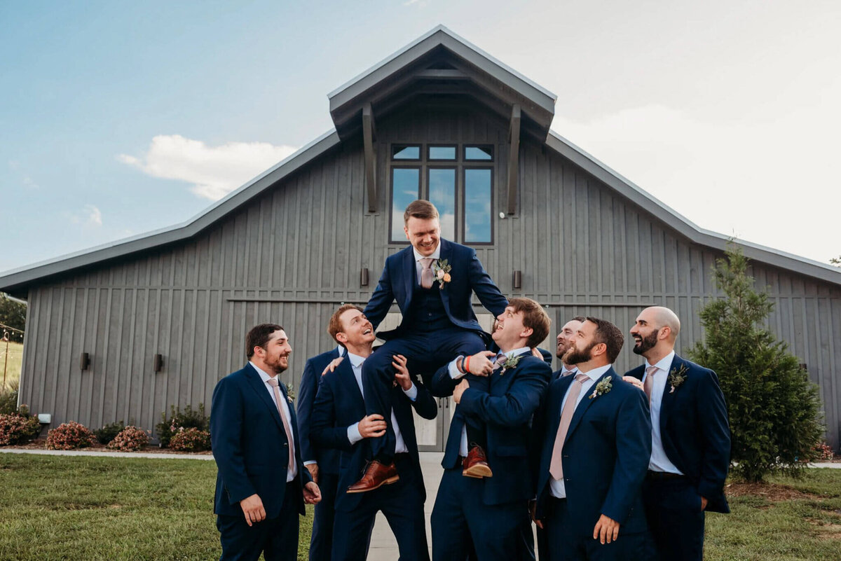 Photo of a groomsmen sitting on the shoulders of his groomsmen and looking at them in front of a barn wedding venue in Tennessee