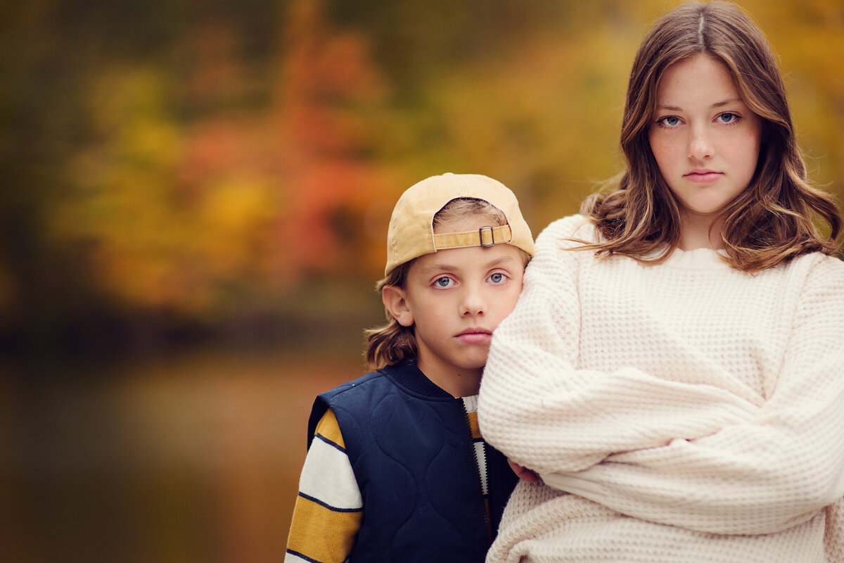 Tween and teen siblings with straight faces and brightly colored fall leaves behind them.