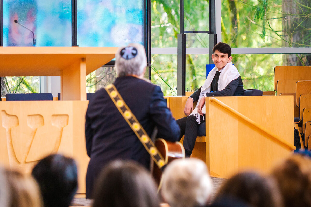 A boy smiles from his chair on the bimah as a man plays guitar