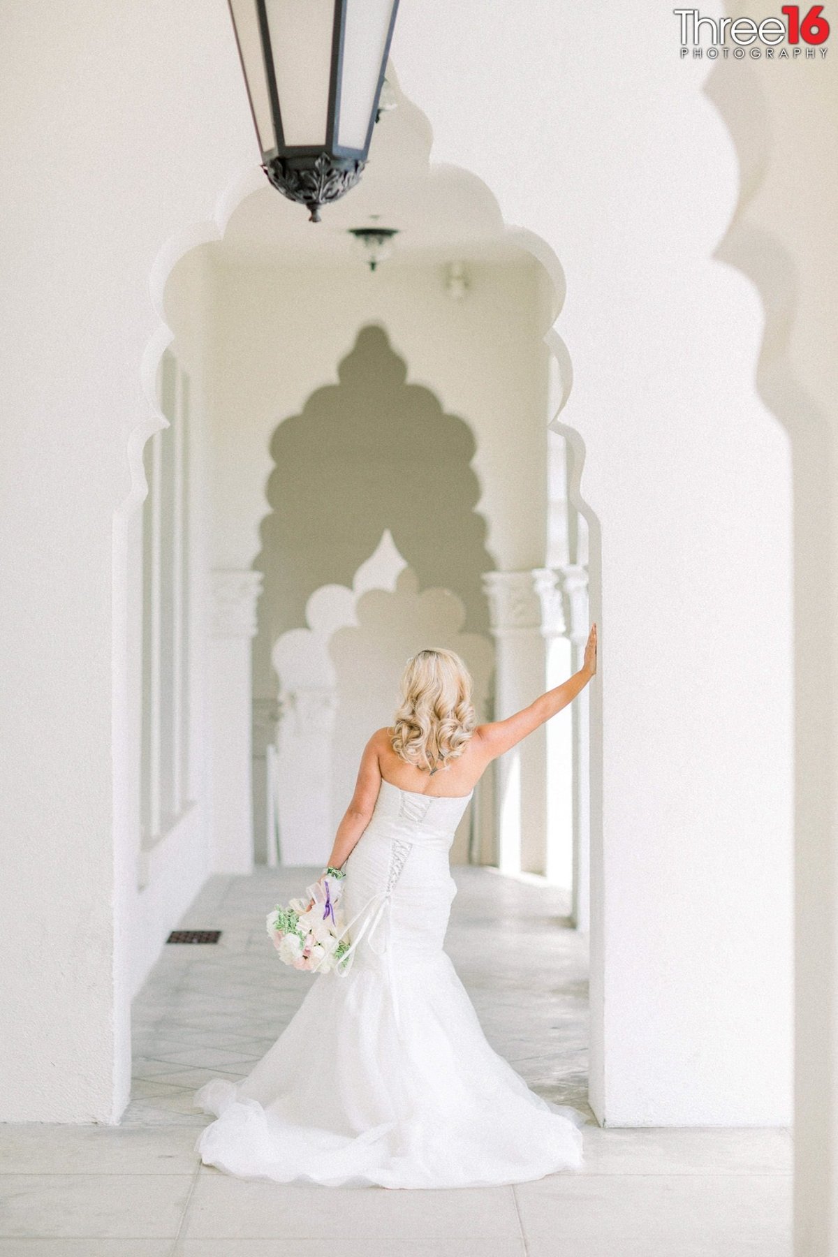 Bride leans against a column facing away from the wedding photographer hold her bouquet down by her side