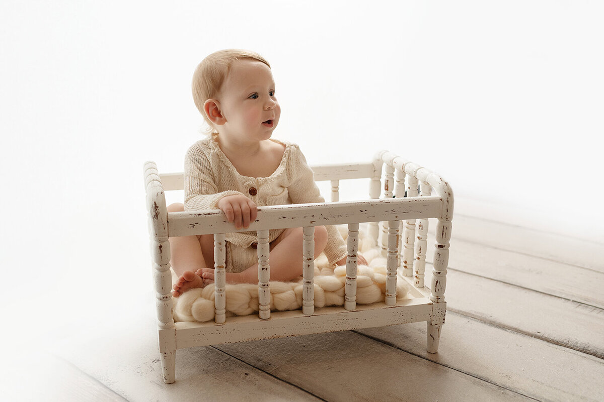 A toddler boy in a white onesie sits in an aged wooden crib in a studio