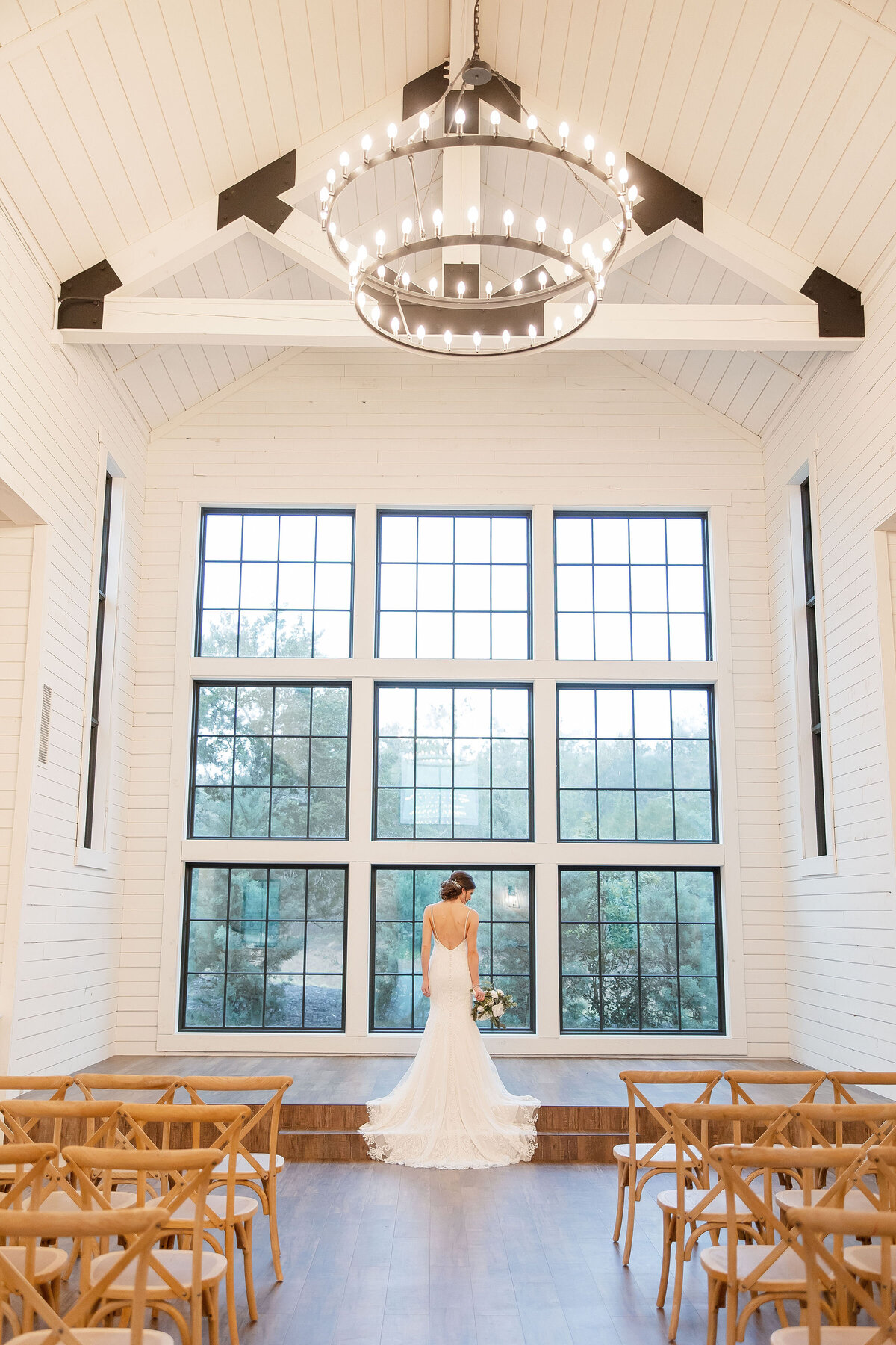 bride stands under chandelier  showing back of dress before 9 large windows at Morgan Creek Barn in Dripping Springs Texas