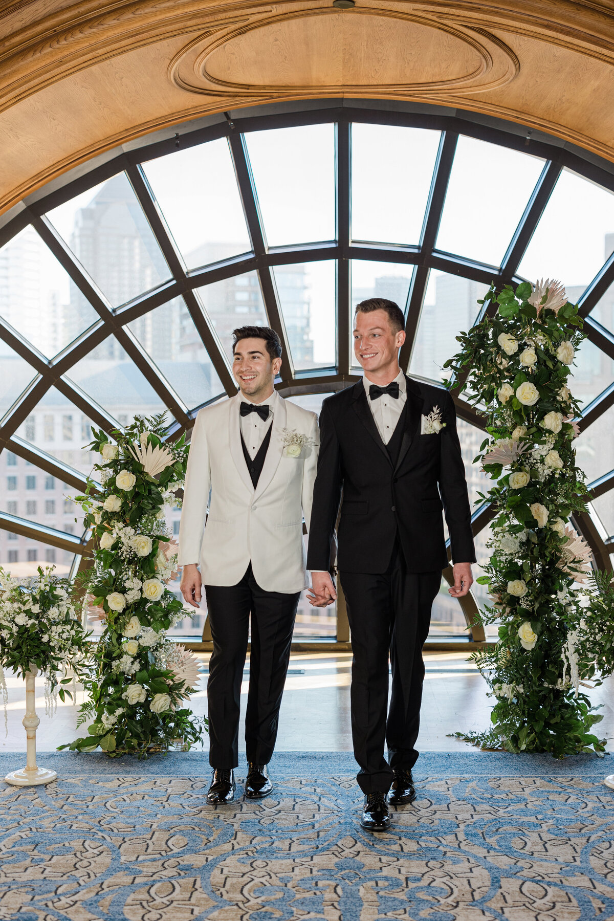 A portrait of two grooms on their wedding day at The Crescent Club in Dallas, Texas. The groom on the left is wearing a tuxedo with a white jacket, black bowtie, and boutonniere. The groom on the left is wearing an all black tuxedo with a bowtie and a boutonniere. They both hold hands while walking down the aisle towards the camera. They are flanked by intricate and large floral arrangements on either side.
