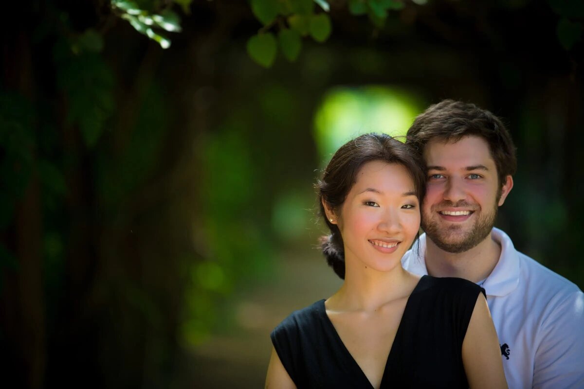 Bright and joyful portrait of a couple, with the woman smiling at the camera and the man smiling at her, framed by lush green foliage