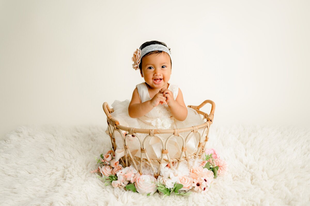 1-year-old girl wearing a headband and a boho style outfit sitting in a basket surrounded by flowers on a cream color rug for her first birthday Milestone session