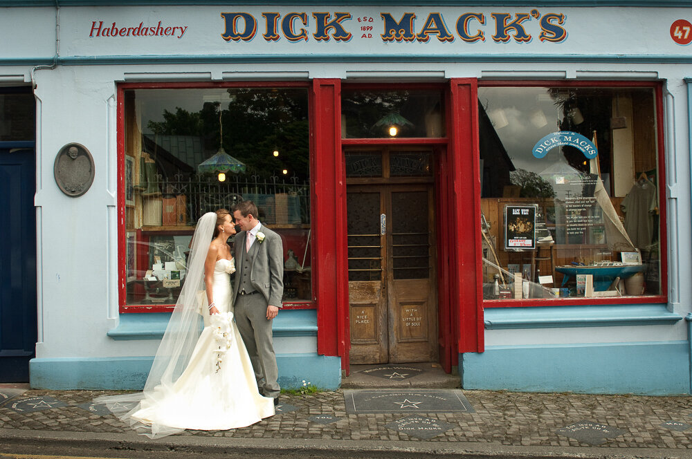 bride wearing a trumpet style wedding dress with corset top with groom wearing a light grey coat tail jacket outside Dick Mack's pub in Dingle