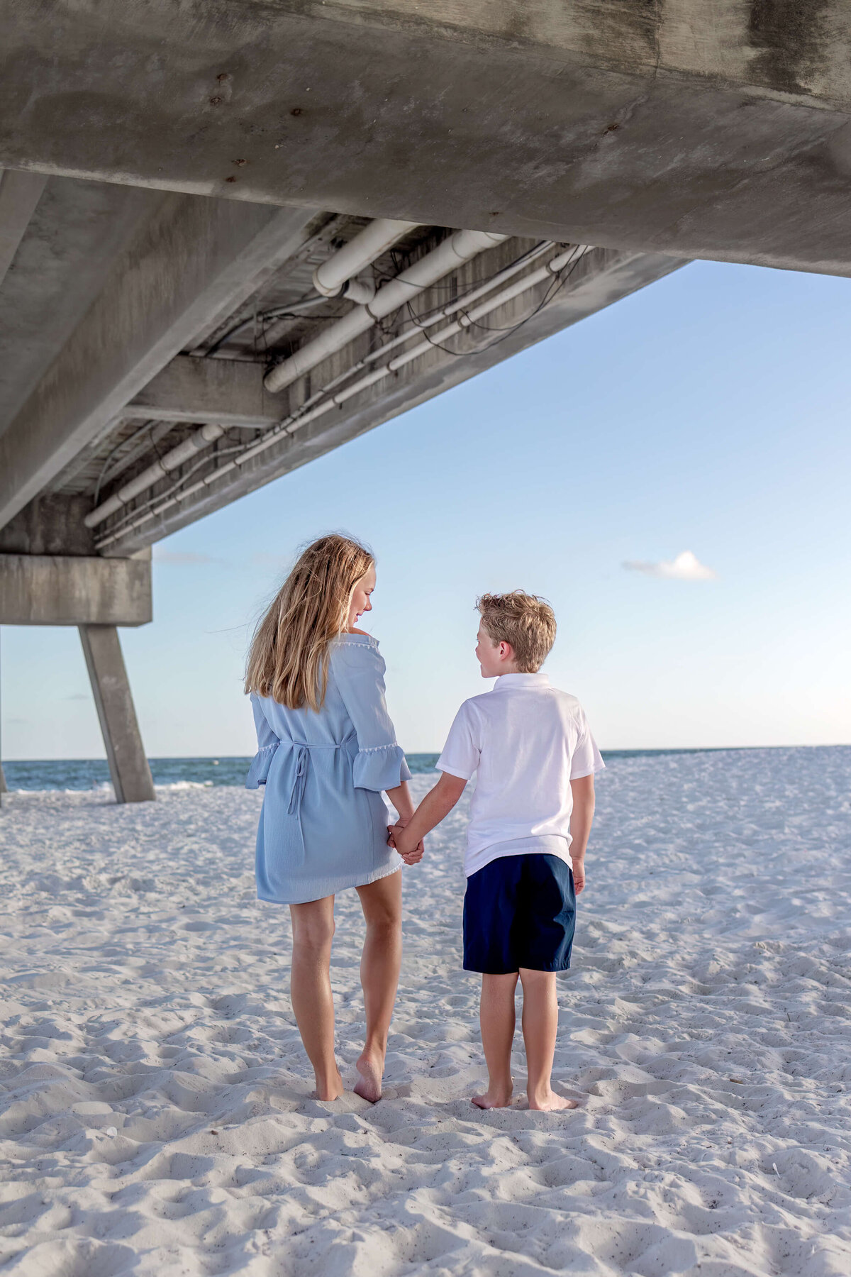 a young boy and girl holding hands loooking at eachother with their bbacks to the camera under a pier next to the ocean