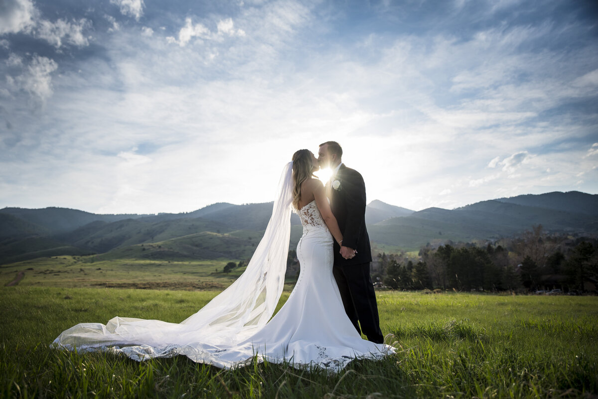 A bride and groom share a kiss in a field at golden hour with the Colorado landscape behind them.