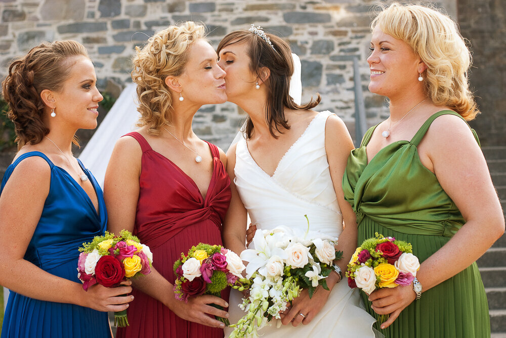 bride with v-neck, a-line, layered tulle dress kissing one of her bridesmaids who are wearing wine, blue and green dresses