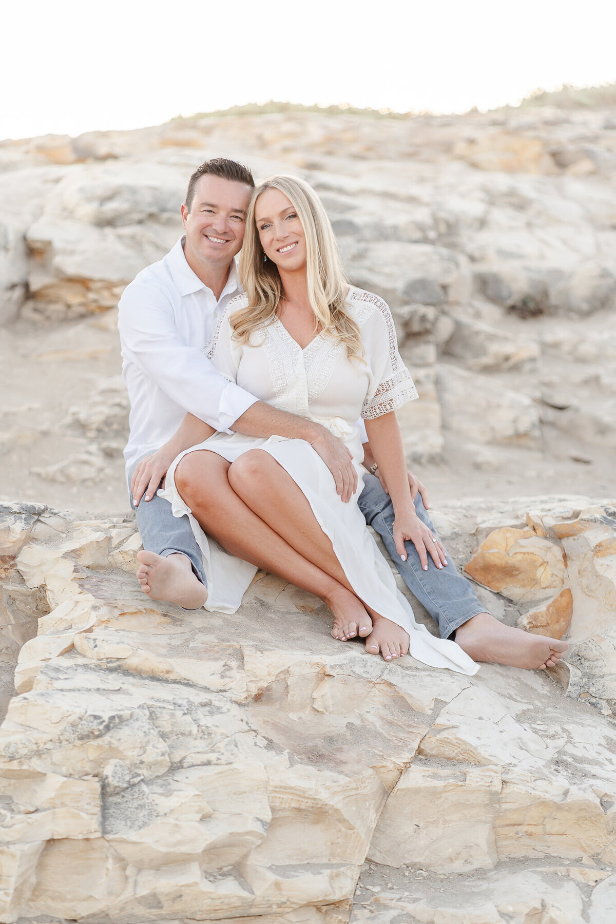 husband and wife wearing white and sitting on a rock at the beach