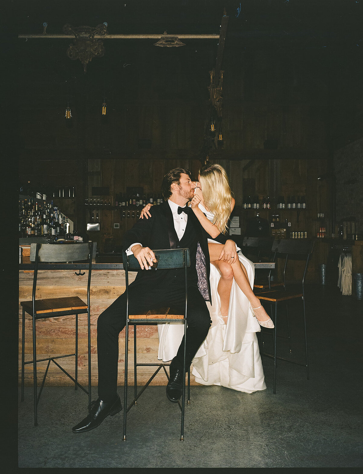 A couple dressed elegantly, a man in a tuxedo and a woman in a long dress, sharing a kiss while sitting at a bar in a dimly-lit rustic room during their Park