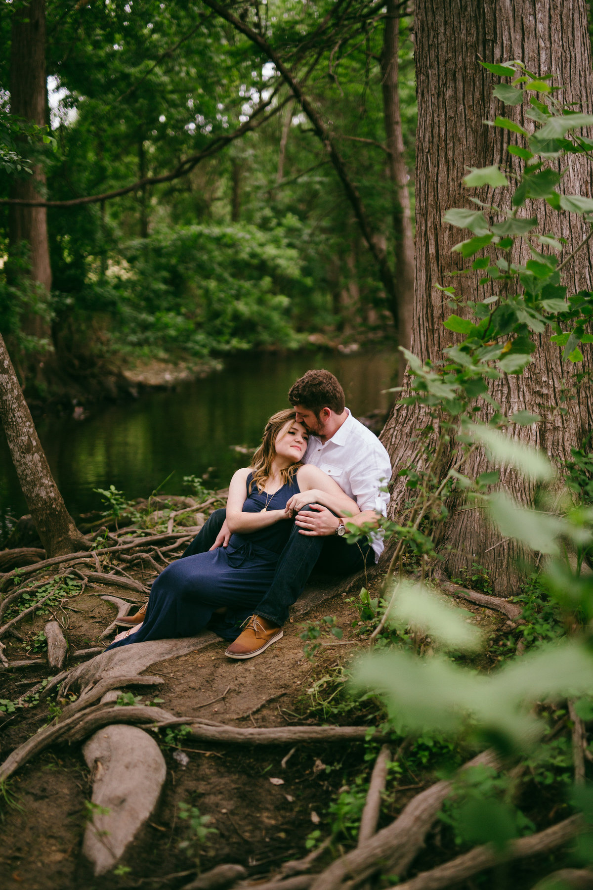 engaged couple sitting under a big tree by a beautiful river. Fiancée is leaning on her fiancé's chest.