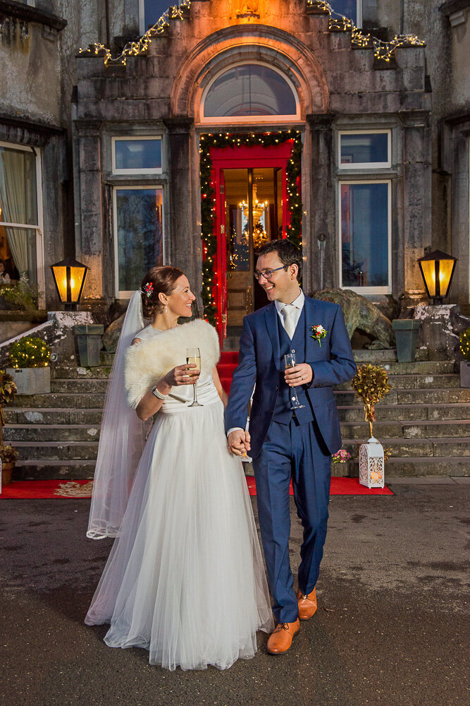 bride in a luxuriant style tulle wedding dress walking with groom in a navy suit and brown shoes, holding champage in front of Ballyseede Castle with Christmas lights