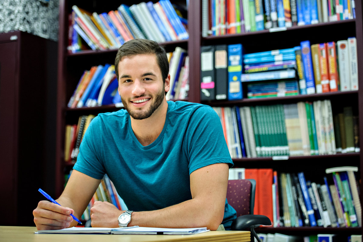 A college student studying at a desk with a bookshelf behind him.
