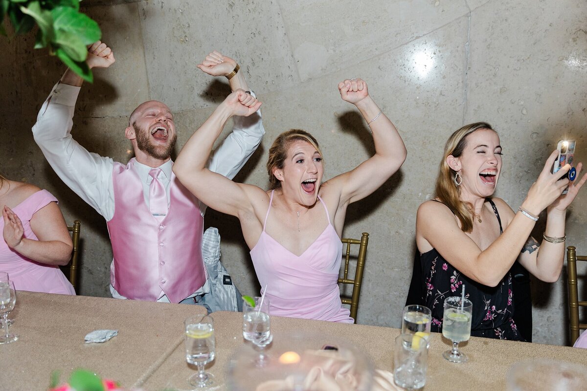 A bride holding up her arms and cheering in joyous celebration during her wedding reception at the Hall of State in Fair Park in Dallas, Texas. The bride is wearing a sleeveless, pink dress. Members of her wedding party sit to her right and are also cheering while other guests sit and celebrate on her left.