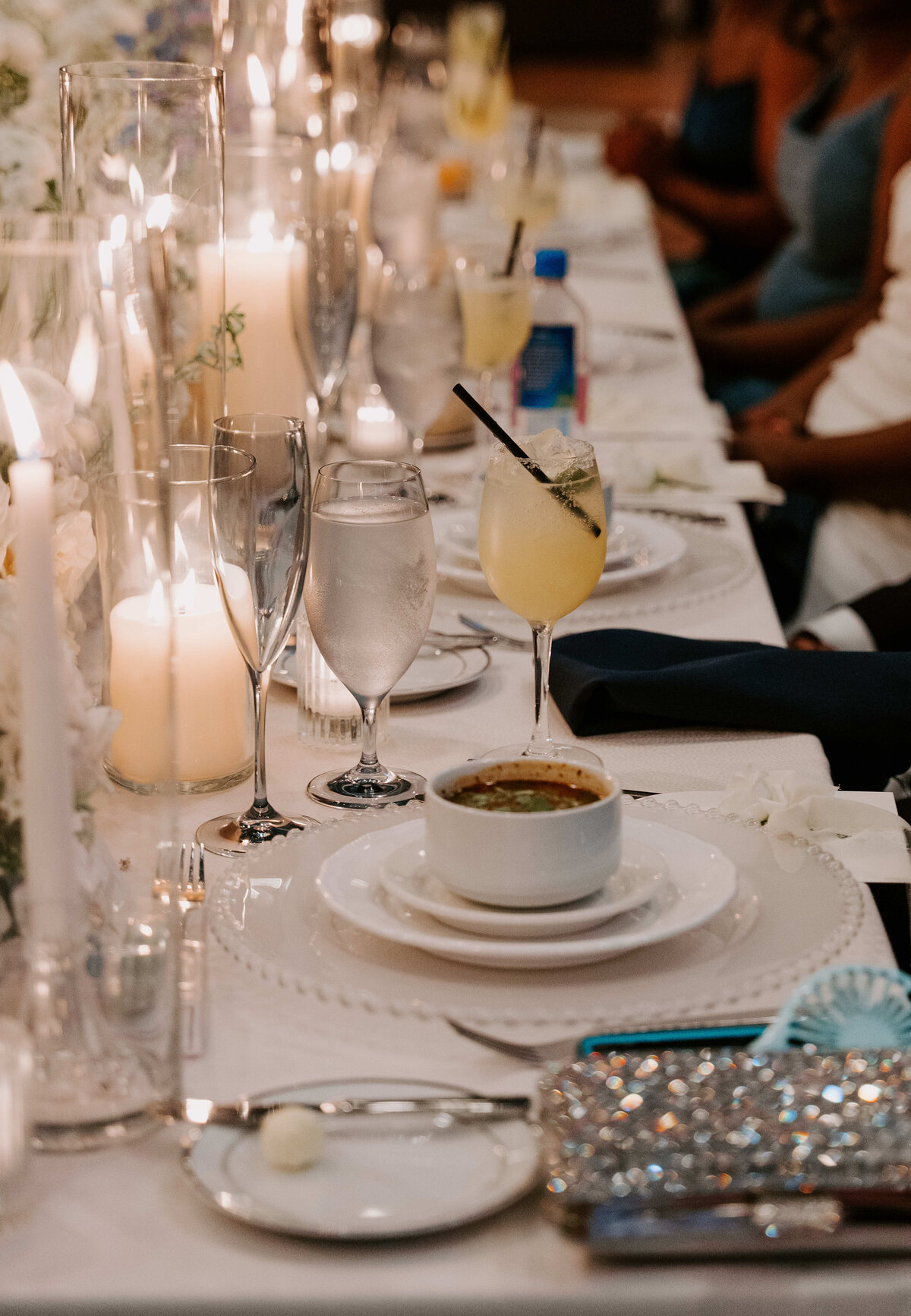Elegant dinner setting along a white tablecloth with candles, champagne glasses, a soup bowl, and a cocktail. Guests in formal attire in background.