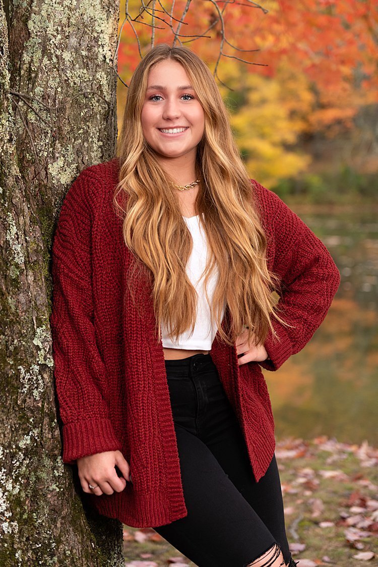 High school senior girl in red sweater leaning on tree with fall leaves