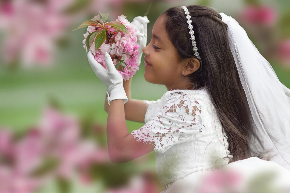 A young girl in a white lace dress smells some pink flowers for a New Jersey Communion Portrait Photographer