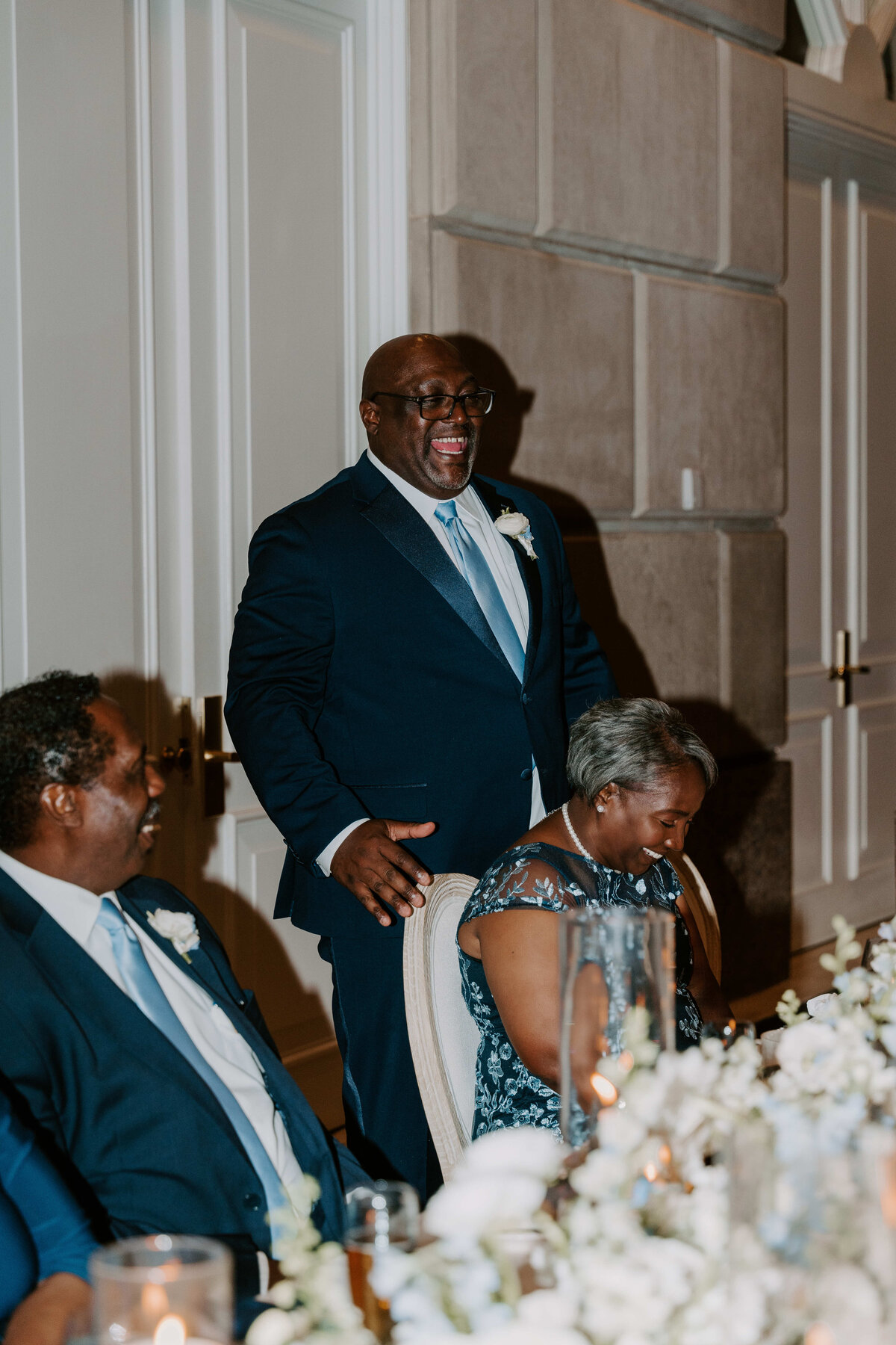 A joyful man in a navy suit stands, laughing heartily at a formal event. Two seated individuals in elegant attire smile. The setting is sophisticated and warm.