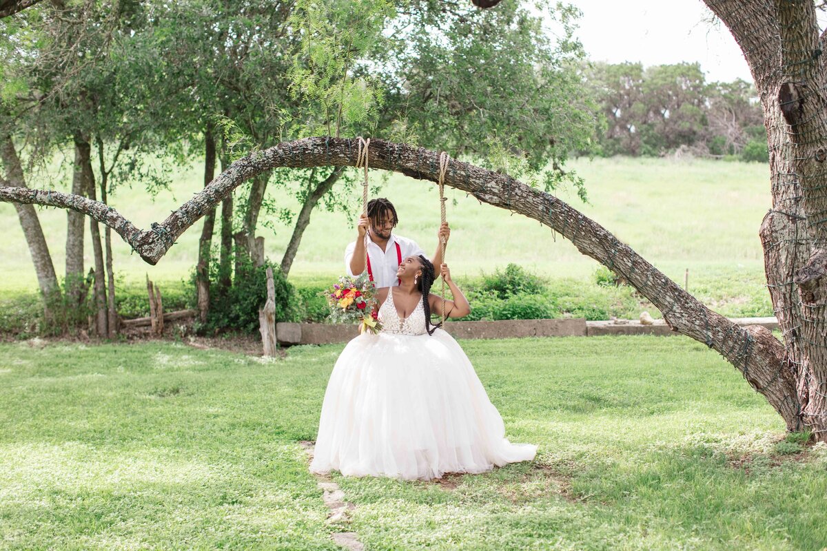 groom stands behind bride on swing holding each side under arched tree at wedding near Austin Texas