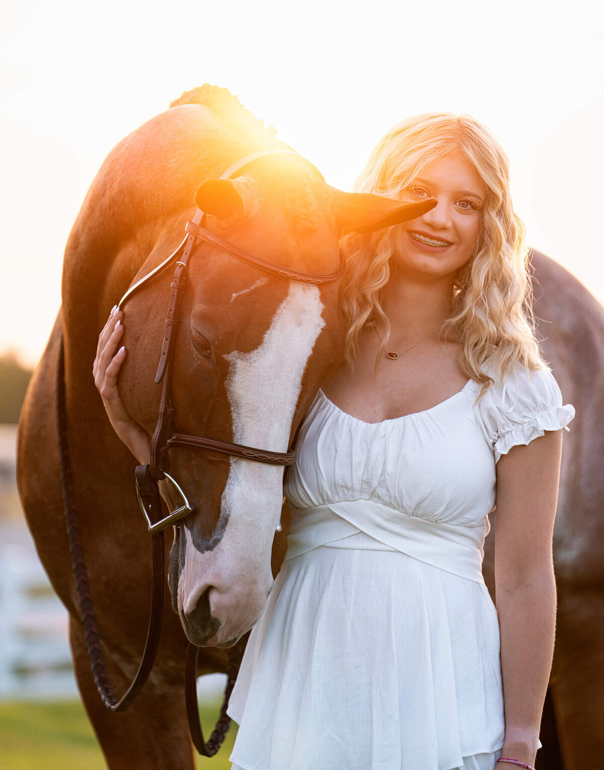 young blonde equestrian in a white jumper suit with her chestnut paint horse nestled into her side. behind them is beautiful white fence, trees, and golden hour sun.