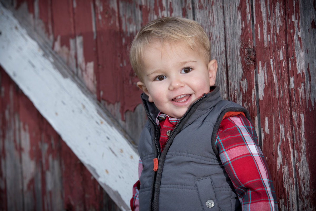 Smiling  toddler in a vest against a historic red barn during a family portrait