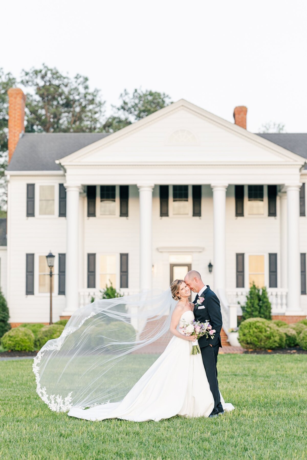 Bride and groom embracing as her veil flys in front of white mansion