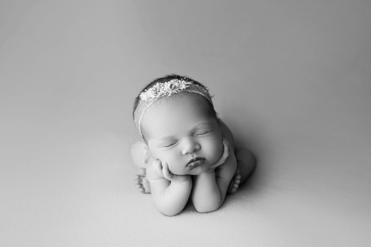 Philadelphia's best newborn photographer, Katie Marshall, captures a black and white image of a newborn baby girl sleeping in froggy pose. Baby is wearing an embellished lace headband and her hands are folded under her chin.