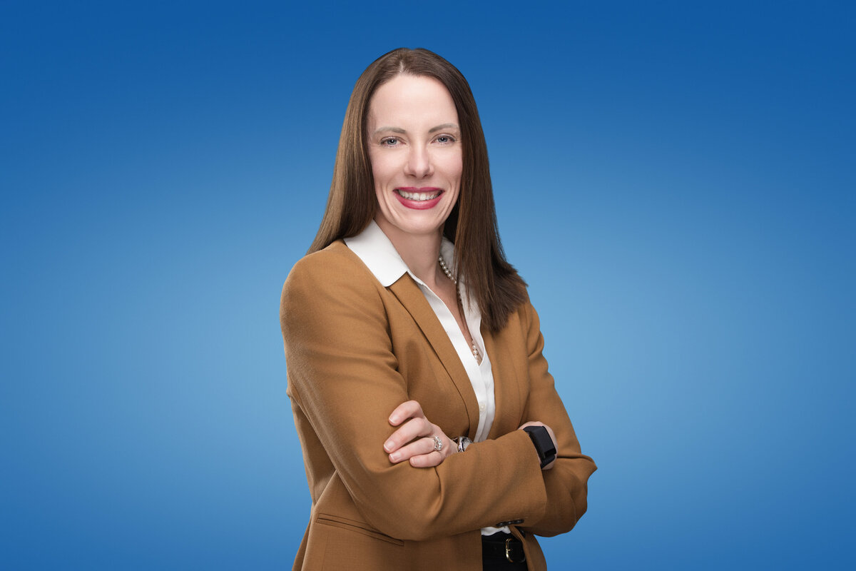 Professional headshot of a confident woman with long, straight brown hair, wearing a camel blazer over a white blouse, accessorized with a pearl necklace. She stands with her arms crossed and offers a warm, approachable smile. The background features a smooth, gradient blue backdrop. Photographed at Janel Lee Photography, a professional headshot and personal branding studio in Cincinnati, Ohio.