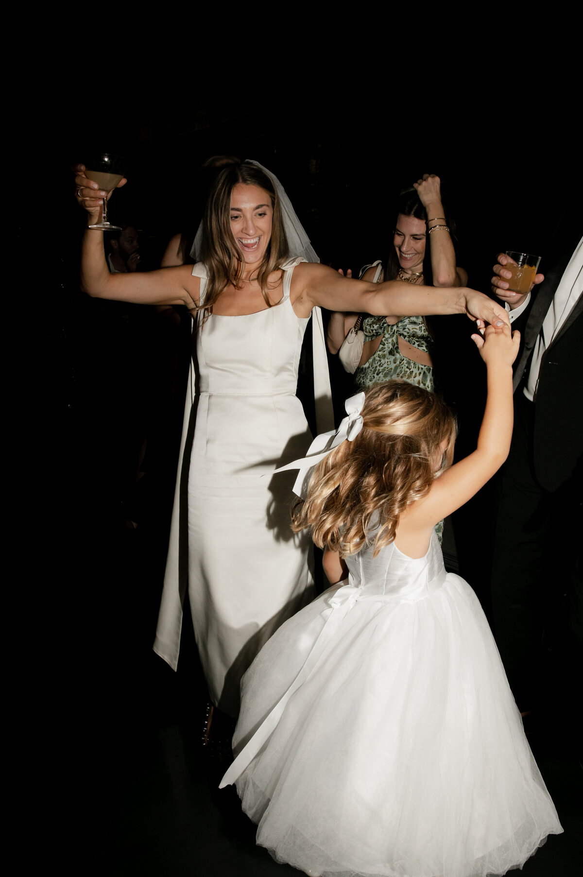 Smiling elegant bride holding wine twirls flower girl on the dance floor while guests dance in the background at Elske Restaurant wedding reception.