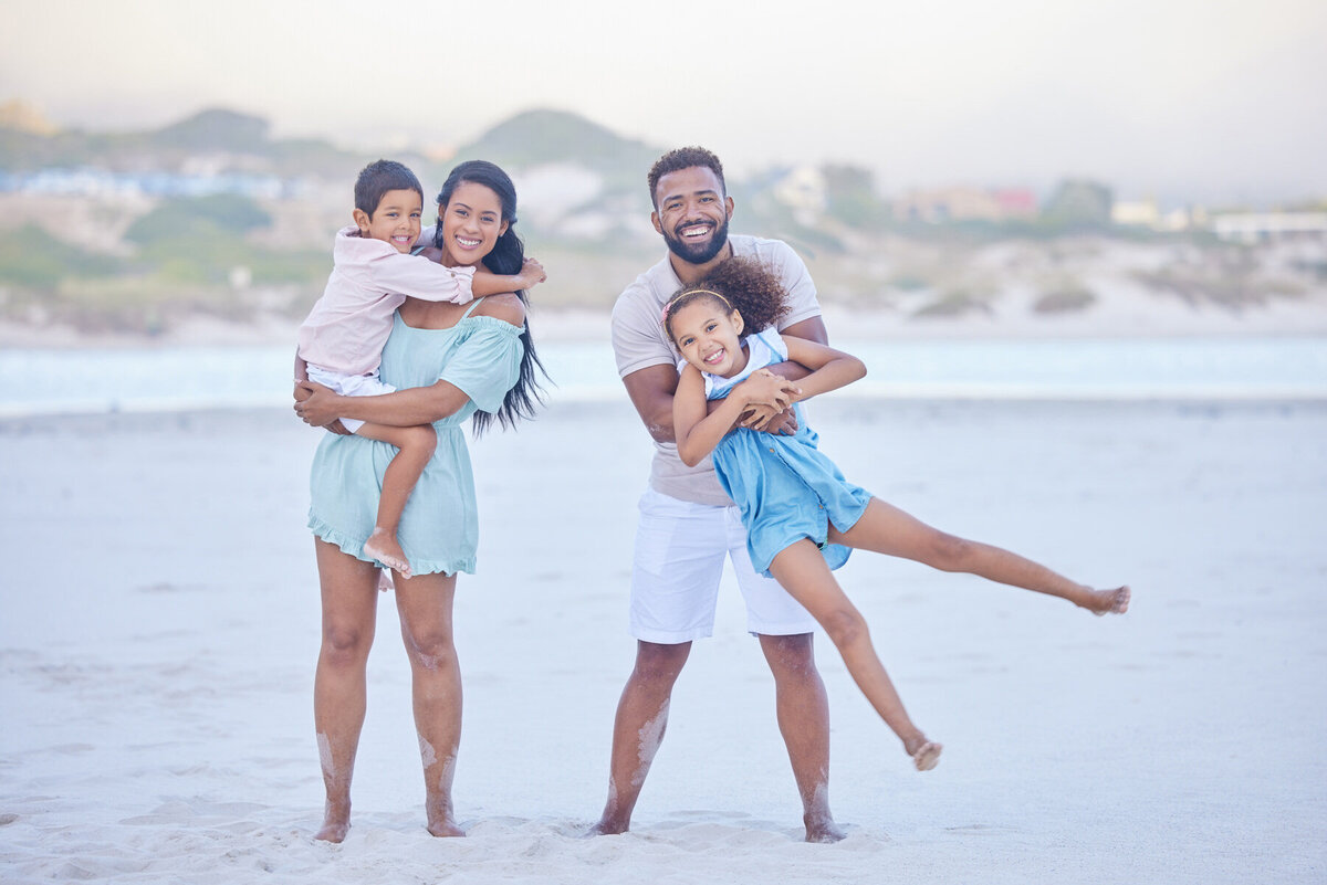 A joyful family of four poses on a beach. The parents are each holding a child; the mother holds the son, and the father holds the daughter. They are all smiling, with a sandy beach and blurred background of hills and buildings.