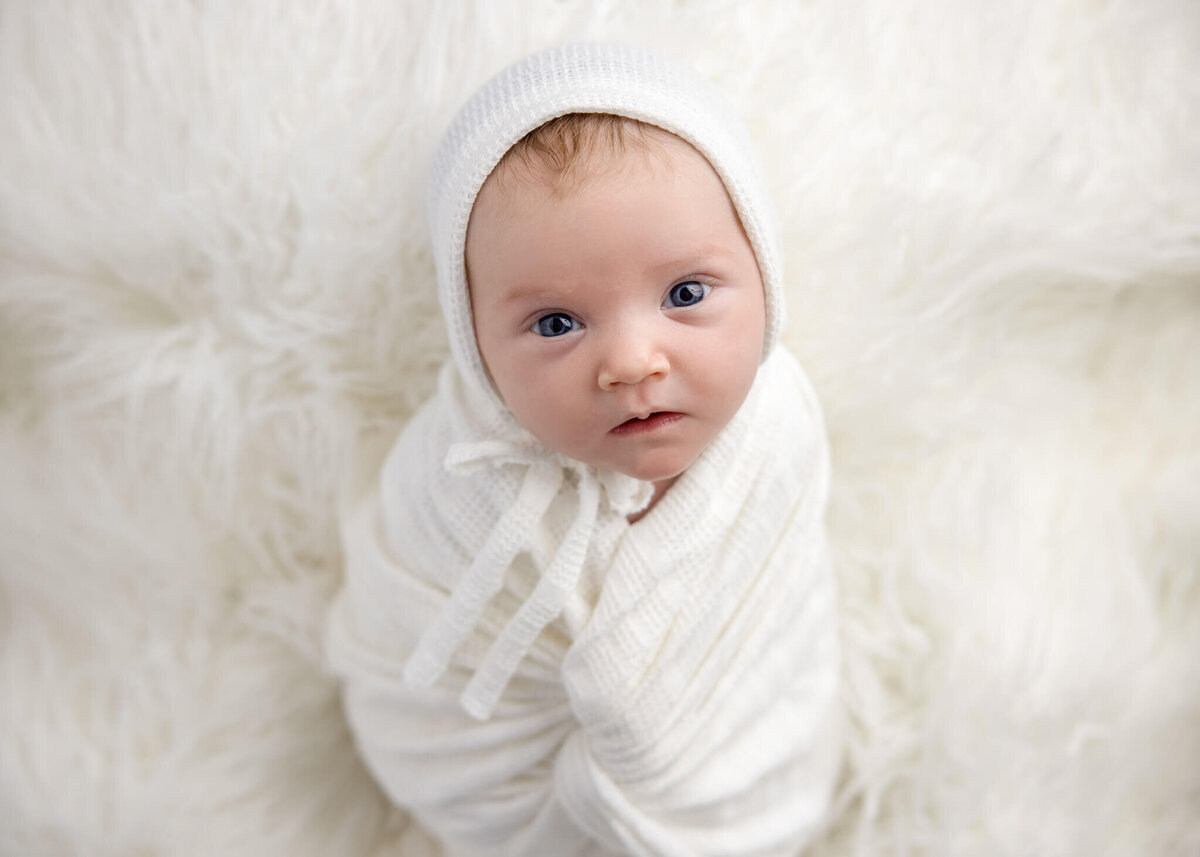 close up of newborn baby face with eyes open wrapped in white fabric and white bonnet