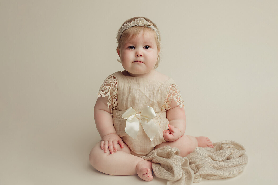 one year old girl wearing a cream lace romper sitting with a cream fabric looking at the camera