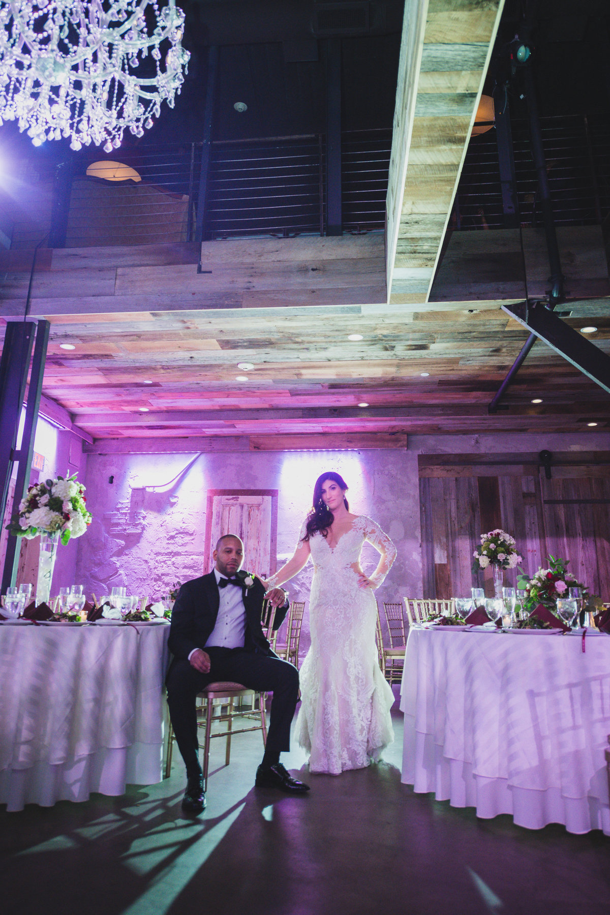 wedding photo of groom sitting with bride standing holding his shoulder in wedding reception at The Loft by Bridgeview