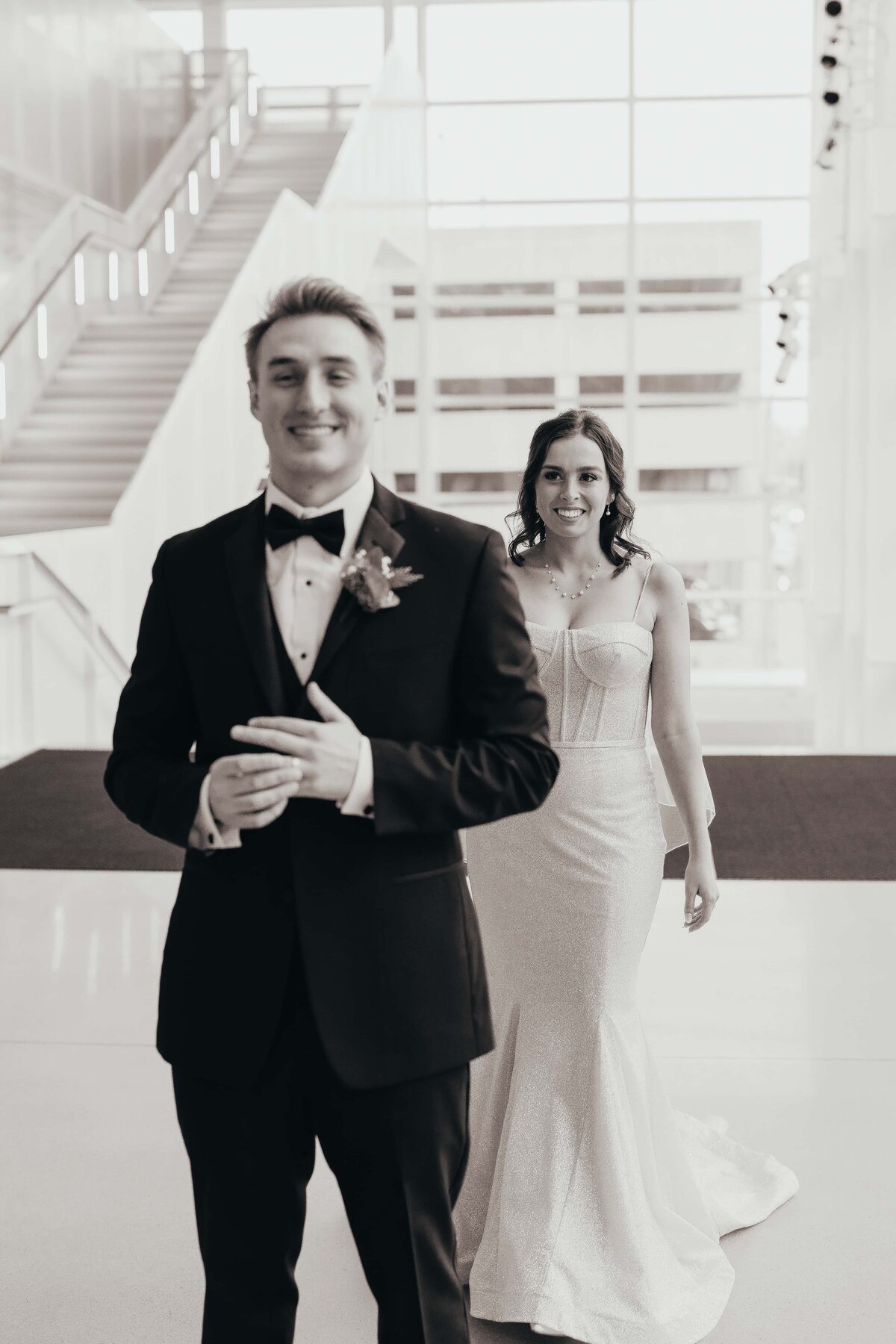 A bride and groom smiling in a modern building with white stairs and large windows at an Iowa wedding.