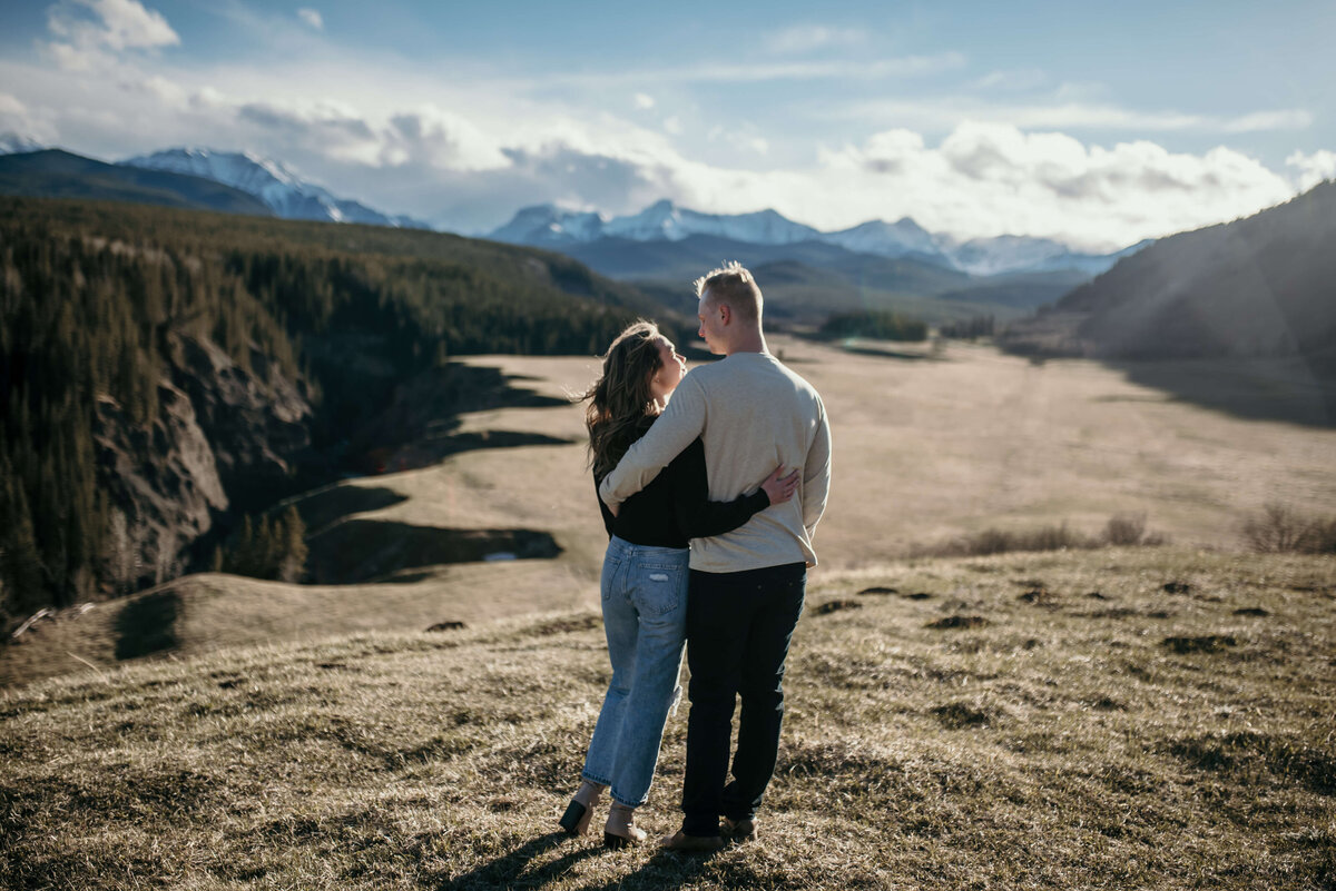 Man and woman with arms crossed over each others backs looking at each other with mountains in the background