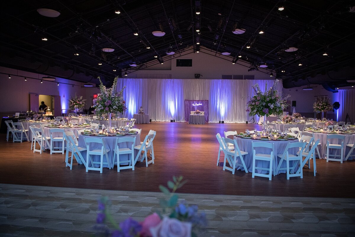 Wedding reception at with round tables covered with purple linens and surrounded by white garden chairs. Tall floral arrangements of purple roses and greenery on clear glass vases sit on each table. The sweetheart table sits in front of a purple sequined backdrop and neon sign. The back wall is covered with white drapery and purple uplights at Noah Liff Opera Center in Nashville.