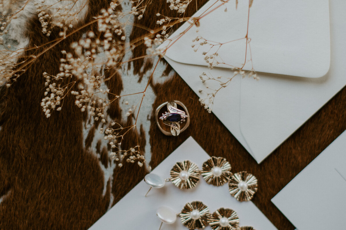 A pair of earrings and wedding bands on white stationery atop cow hide with dried flower stems.