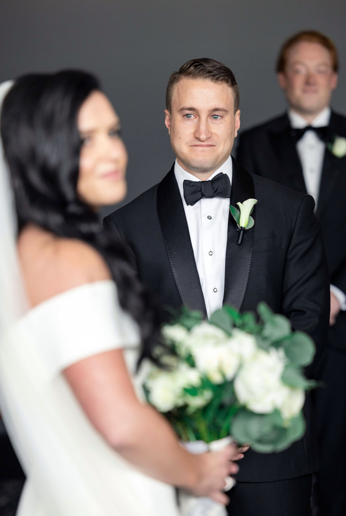 A bride and groom stand together during a wedding ceremony. The groom, in a black tuxedo, looks at the bride, who holds a bouquet of white flowers. Another person in a tuxedo stands in the background.