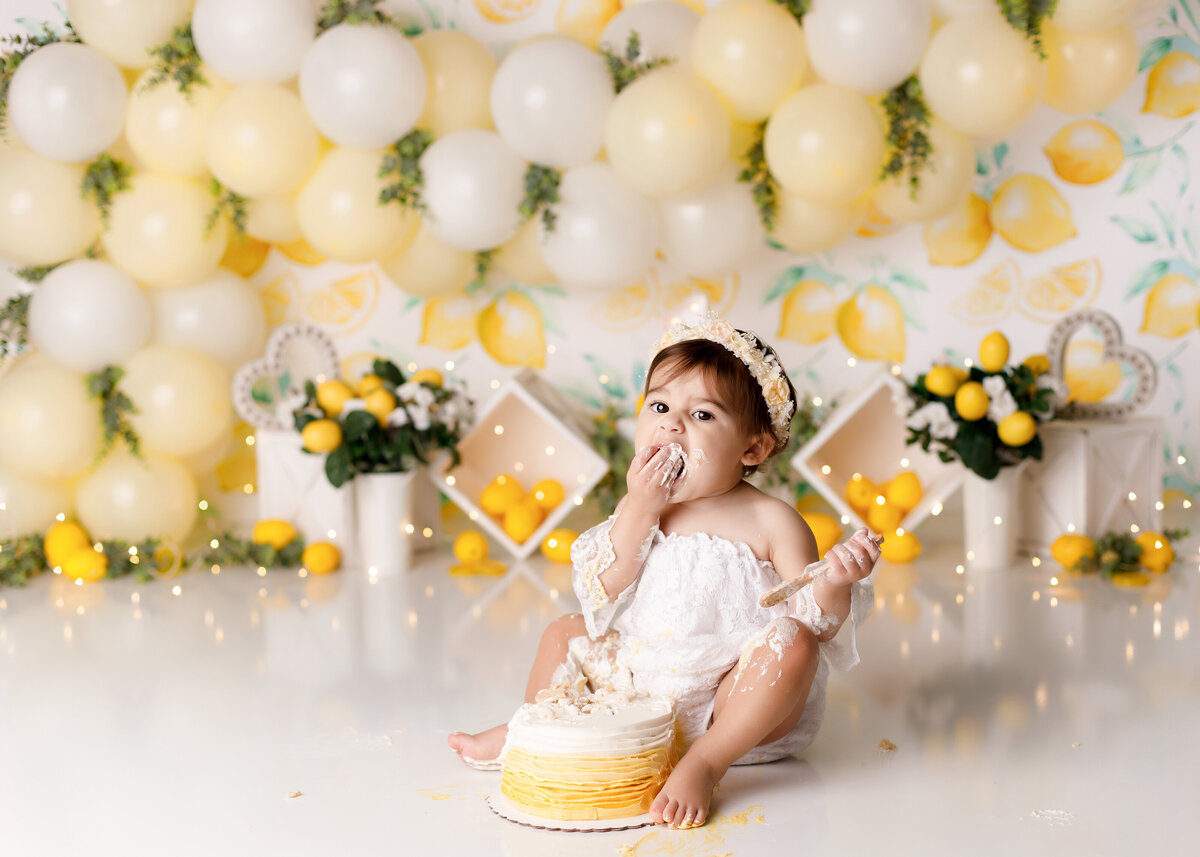 Lemon themed cake smash at West Palm Beach newborn and cake smash photographer. Baby in white lace off the shoulder onesie sitting in front of a yellow ombre cake. She has cake in both fists and taking a bite looking at the camera. There is a lemon-print backdrop, lemons, and lemon branches and a yellow and white balloon arch.