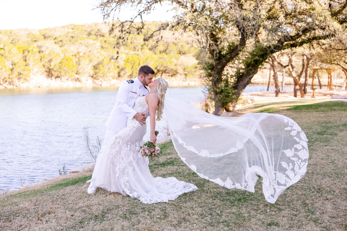 groom in military uniform dips bride in kiss by water with veil flowing at The Preserve at Canyon Lake Texas wedding