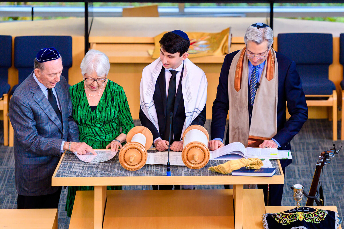 A teenage boy reads from the torah at the bimah with a rabbi and his grandparents