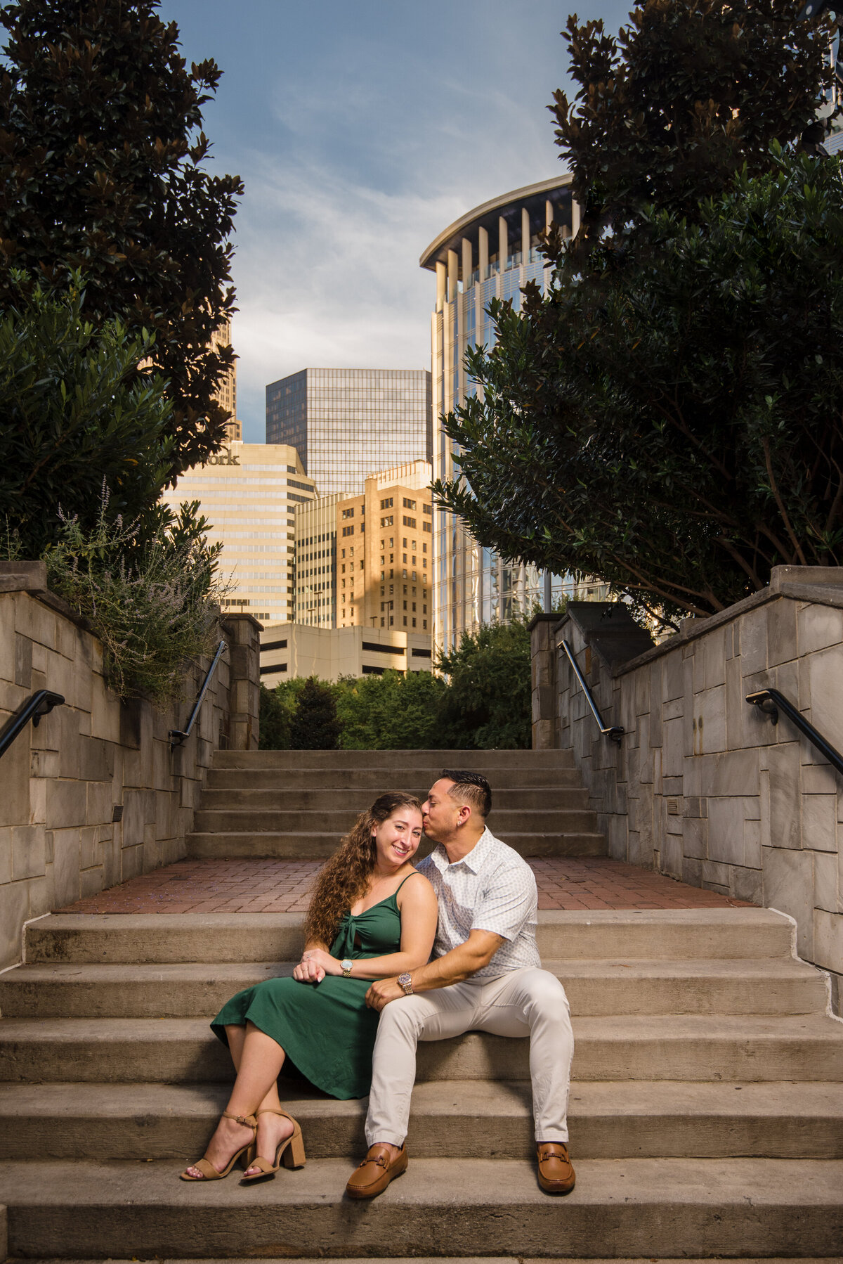 A-couple-seated-on-the-steps-in-Romare-Bearden-park-the-man-kissing-the-woman-on-her-temple-as-she-smiles-and-looks-at-the-camera-with-Charlotte-city-buildings-in-the-background