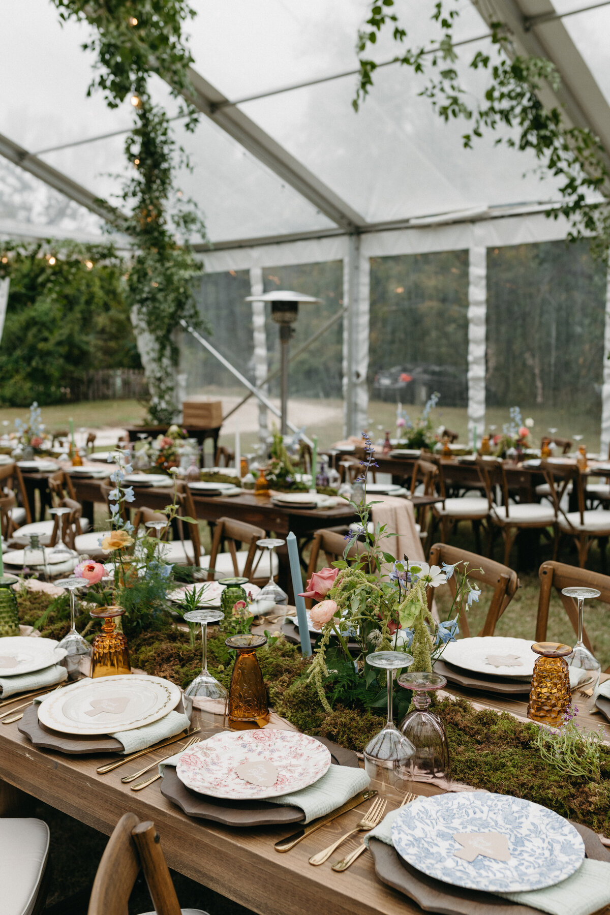 Whimsical garden-inspired wedding reception inside a clear tent, featuring long wooden tables decorated with moss table runners, pastel taper candles, wildflower-style floral centerpieces, and mismatched vintage plates. Hanging greenery installations and soft natural lighting create a romantic outdoor wedding atmosphere in Northwest Arkansas.