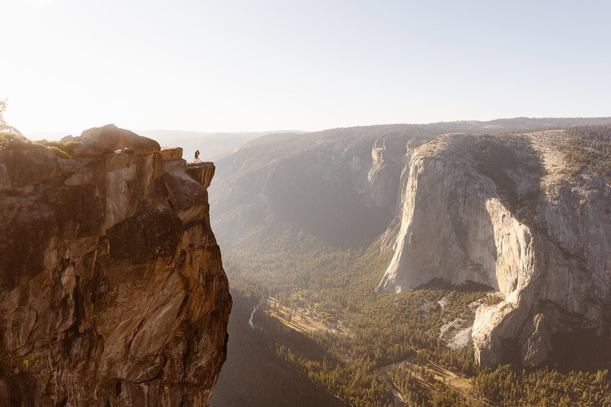 A couple stands at the edge of a dramatic cliff at Taft Point in Yosemite National Park, overlooking the valley far below with El Capitan in the distance.