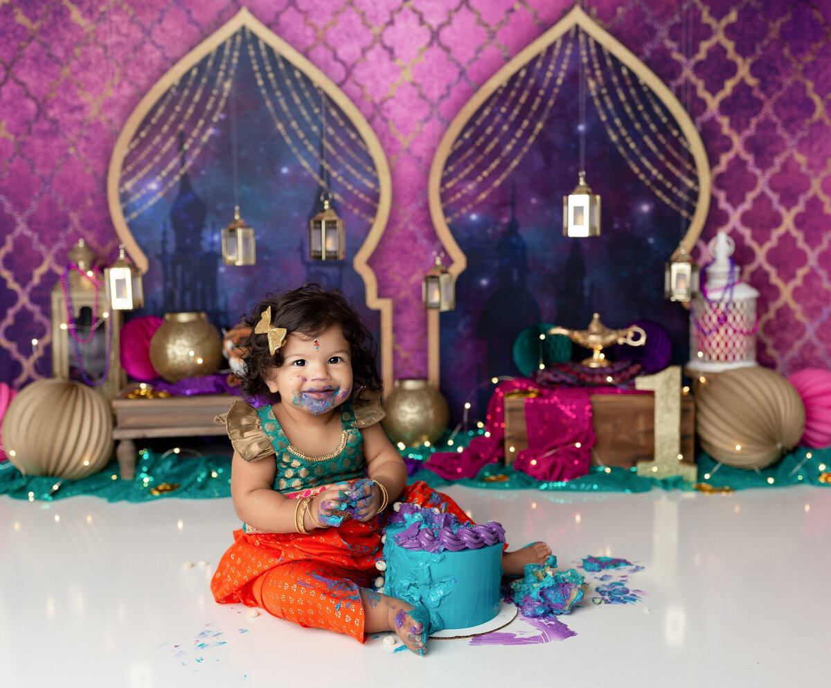 Princess Jasmine Aladdin themed cake smash at West Palm Beach photography studio.  Baby girl is wearing a Jasmine-inspired outfit  with a teal and purple cake smashed between her legs. She has icing on her face, hands, and feet. Behind her, there is a moroccan arch backdrop, and Aladdin's lamp with various shades of magenta, purple, blue, and green.