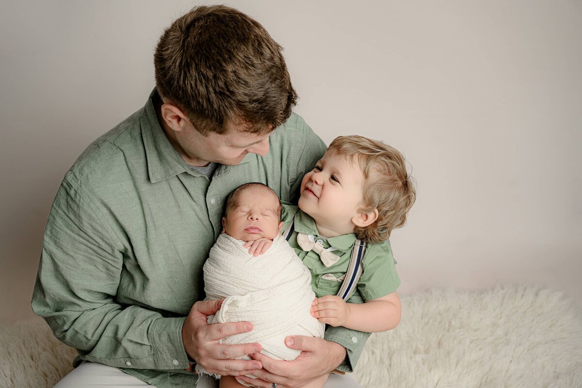 father holds his toddler boy while his newborn son sits in his lap wrapped in white