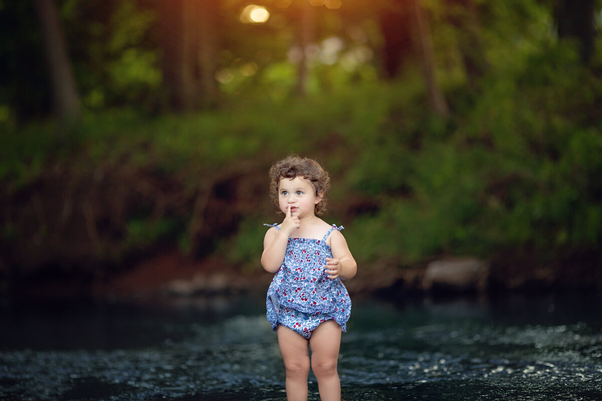 A toddler girl wearing a bathing suit wades through a shallow running river in a forest at sunset