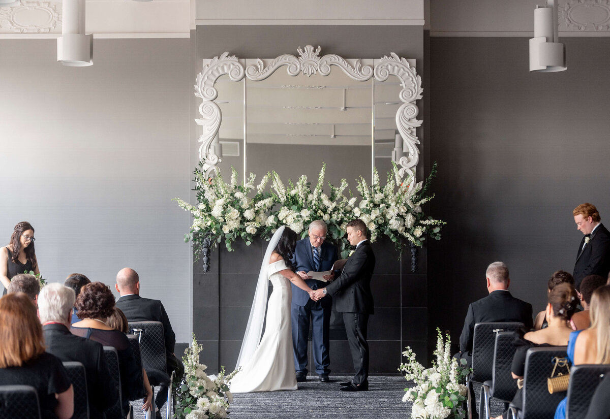 A couple stands at a wedding altar adorned with white flowers and a decorative mirror. They hold hands while exchanging vows in front of an officiant. Guests are seated on either side, watching the ceremony.